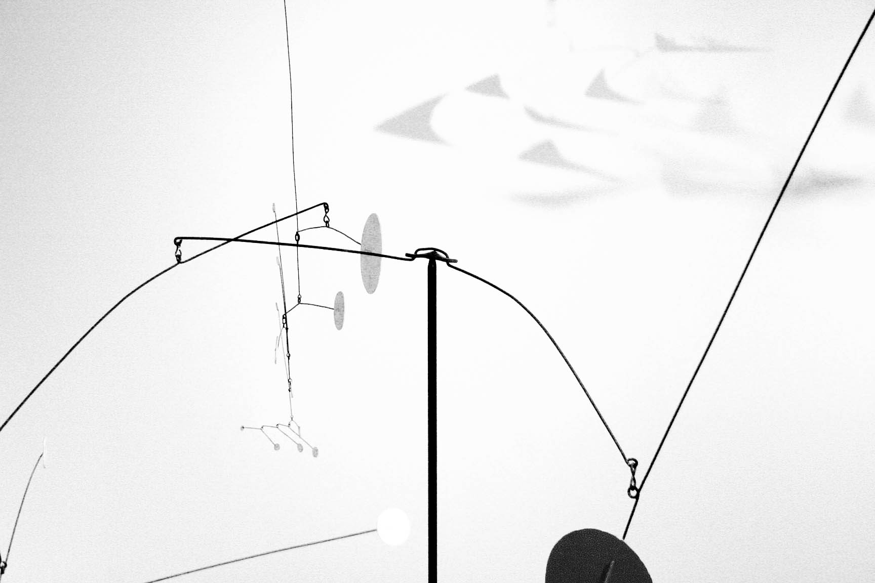 Black and white close-up of a hanging mobile sculpture with thin wires and shapes against a plain background, quiet and minimal mood
